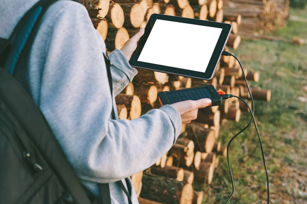 Mock-up of a tablet in the hands of a girl with a power bank charger. Against the background of folded timber, round wood - Φωτογραφία, εικόνα