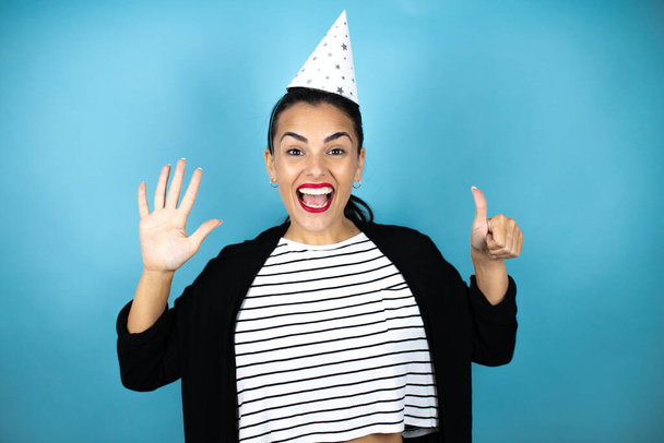 Jovem mulher bonita usando um chapéu de aniversário sobre fundo azul insolado mostrando e apontando para cima com os dedos número seis enquanto sorri confiante e feliz - Foto, Imagem