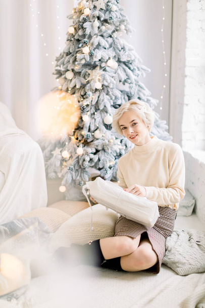 Happy positive Caucasian blond woman in trendy sweater and skirt, sitting indoors on the floor with cozy Christmas decorations, pillows, opening a Christmas box. Christmas tree on the background - Фото, изображение