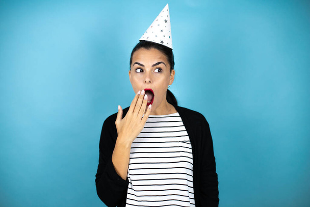 Jovem mulher bonita vestindo um chapéu de aniversário sobre insolado fundo azul surpreendido cobrindo sua boca - Foto, Imagem