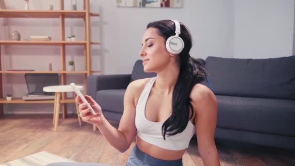 Smiling sportswoman using smartphone and headphones at home  - Footage, Video
