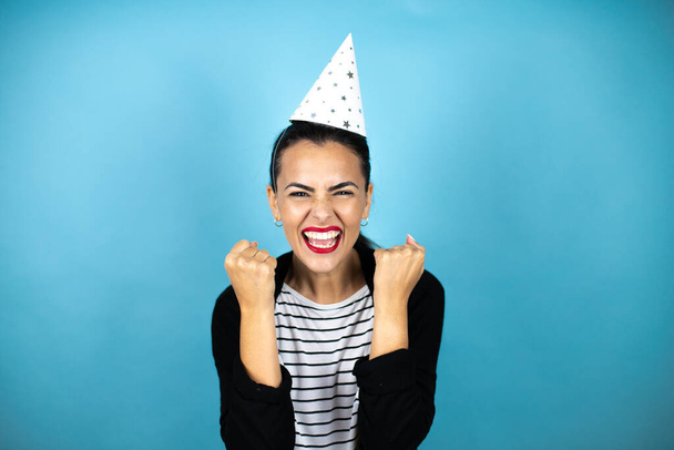 Young beautiful woman wearing a birthday hat over insolated blue background very happy and excited making winner gesture with raised arms, smiling and screaming for success. - Фото, изображение