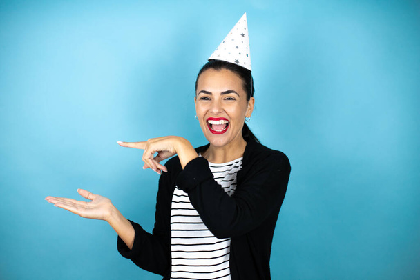 Young beautiful woman wearing a birthday hat over insolated blue background surprised, showing and pointing something that is on her hand - Фото, изображение
