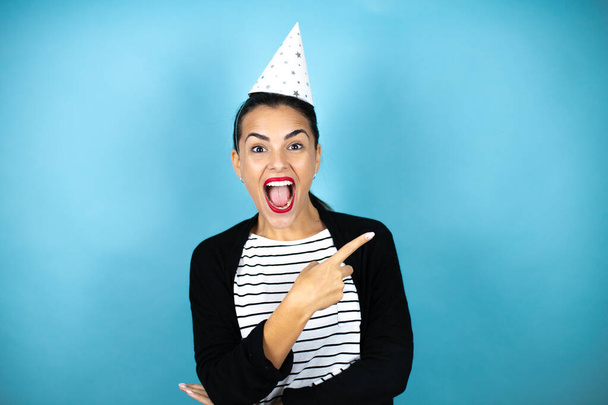 jonge mooie vrouw het dragen van een verjaardag hoed over geïsoleerde blauwe achtergrond glimlachen en wijzen met hand en vinger naar de zijkant - Foto, afbeelding