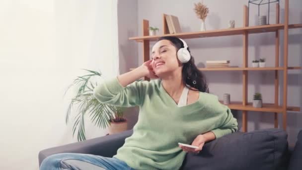 Χαρούμενη γυναίκα που χρησιμοποιεί ακουστικά και smartphone στον καναπέ στο σπίτι  - Πλάνα, βίντεο