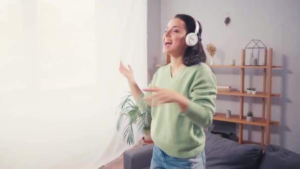 Χαμογελαστή γυναίκα με ακουστικά που χορεύει και τραγουδάει στο σπίτι  - Πλάνα, βίντεο