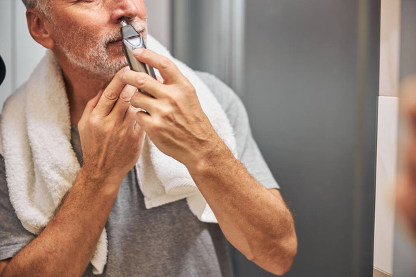 Homem idoso com um aparador de cabelo do nariz em pé perto do espelho - Foto, Imagem