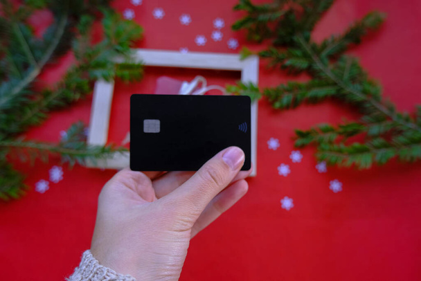 henkilö, jolla on musta luottokortti punaisella taustalla lahjapakkauksella ja joulukoristeella. Joulumyynti, ostokset konsepti - Valokuva, kuva