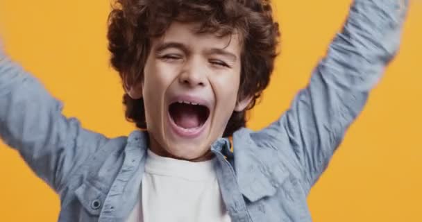Podniecony młody chłopiec cieszący się wygraną, podnosząc ręce i krzycząc, pomarańczowe tło studio, zbliżenie - Materiał filmowy, wideo