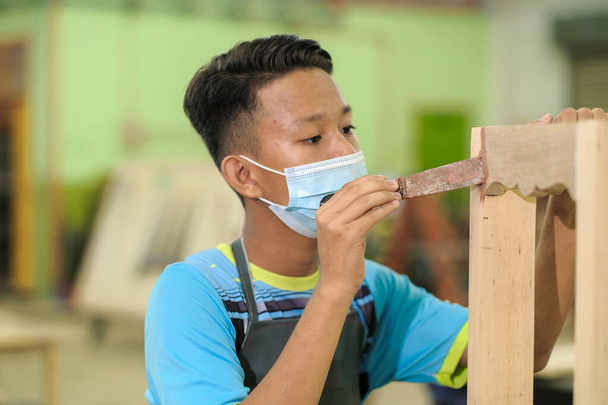 Jovem carpinteiro em roupas de trabalho e máscara facial usando enchimento de madeira no trabalho de acabamento para mesa de console . - Foto, Imagem