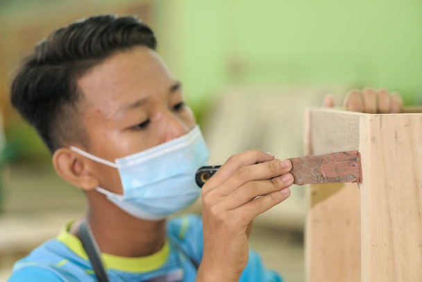 Giovane falegname in abiti da lavoro e maschera facciale utilizzando riempitivo in legno nel lavoro di finitura per consolle . - Foto, immagini