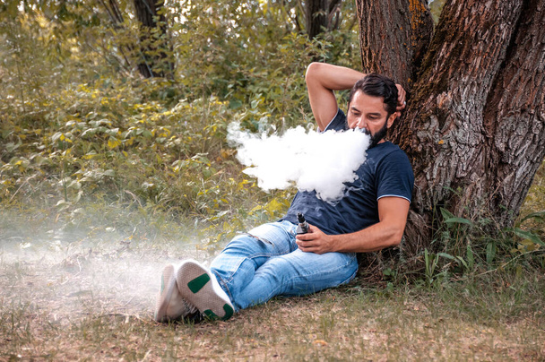 Stilvoller bärtiger Raucher sprengt einem Paar eine E-Zigarette auf dem Waldboden in die Luft. Elektronische Zigarette. - Foto, Bild