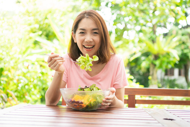 庭の横にある家のバルコニーに座って健康的な緑のサラダを食べる幸せなアジアの健康な中年の女性。健康管理と栄養価の高い食品の概念 - 写真・画像