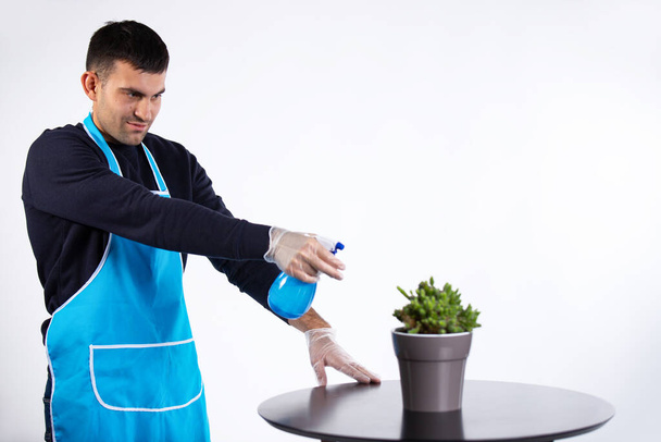 Een man giet water op een indoor bloem uit een spuitfles thuis. Huishoudelijk werk en genderstereotype concept. Foto op witte achtergrond met lege ruimte. Hoge kwaliteit foto - Foto, afbeelding