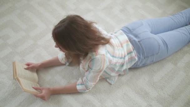 Krásná mladá žena čtení knihy při ležení na podlaze doma - Záběry, video