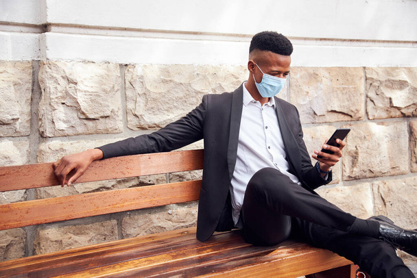 Empresario con máscara sentada al aire libre en el banco con teléfono almorzando durante la pandemia - Foto, imagen