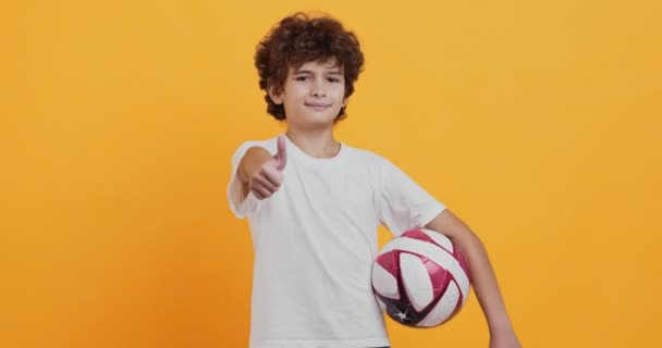 Pequeño jugador de fútbol de pie con pelota de fútbol y gesto pulgar hacia arriba, fondo de estudio naranja - Metraje, vídeo