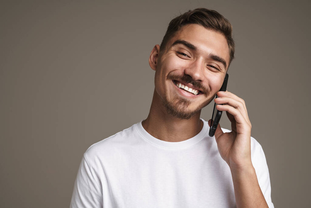 Image of joyful unshaven guy smiling and talking on cellphone isolated over grey background - Фото, зображення
