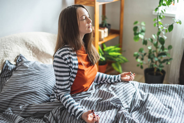 A young woman in pajamas is sitting on the bed in the morning in the lotus position and meditating. Concept of mood for a good day. - Фото, зображення