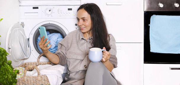 A young woman is resting with a mug of tea and a phone in her hands, she is sitting on the floor next to a pile of laundry. Mom does household chores. - Fotografie, Obrázek