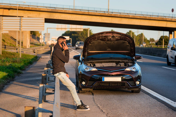 Giovane uomo in piedi vicino a una macchina rotta e chiedendo aiuto o assistenza. Chiamata al meccanico auto per la riparazione auto. - Foto, immagini