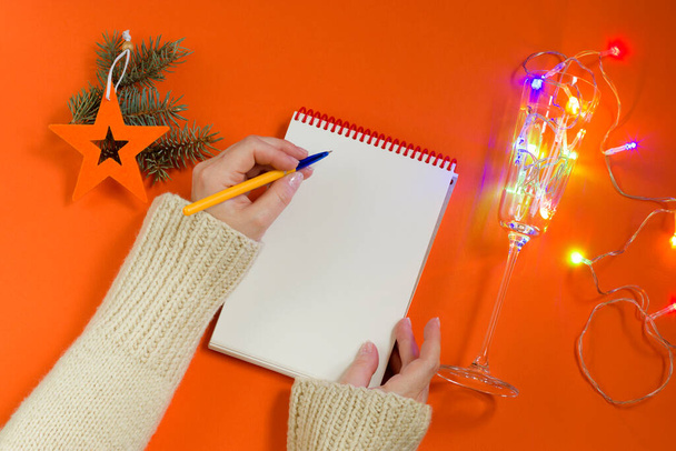 Composición navideña: Adornos del árbol de Navidad, guirnalda, cuaderno y bolígrafo sobre fondo naranja. Una mano femenina escribe los objetivos o planes de Año Nuevo en un cuaderno. Piso tendido, vista superior. - Foto, imagen