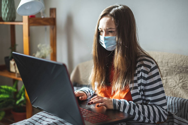Young girl in a protective mask is sitting on a bed at home and using a laptop. Concept of distance learning, remote work, online communication in the pandemic. - Photo, Image