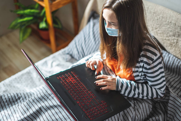 Chica joven con una máscara protectora está sentado en una cama en casa y el uso de un ordenador portátil. Concepto de aprendizaje a distancia, trabajo a distancia, comunicación en línea en la pandemia. - Foto, imagen