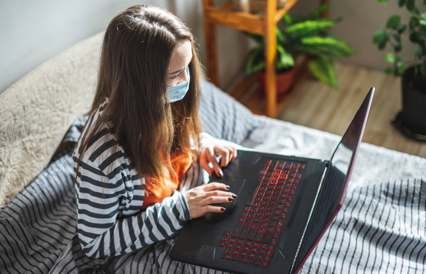 Young girl in a protective mask is sitting on a bed at home and using a laptop. Concept of distance learning, remote work, online communication in the pandemic. - Foto, imagen