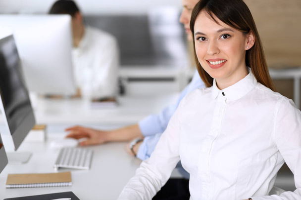 Cheerful smiling asian businesswoman headshot at work in modern office. Casual dressed female entrepreneur using pc computer while sitting with diverse colleagues at the background. Multi ethnic - Фото, изображение