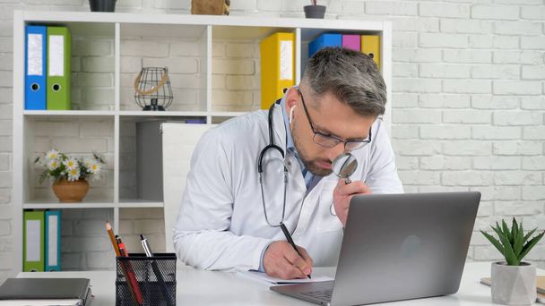 Doktor v nemocniční kanceláři píše diagnózu v notebooku vypadá lupa - Fotografie, Obrázek