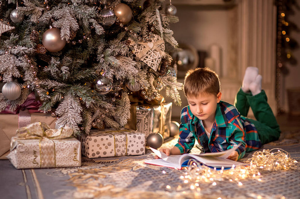 A fiú a padlón fekszik egy díszített karácsonyfa mellett. Egy könyvet olvasok karácsonyi történetekkel. Kellemes téli esték otthon. Fogalom a karácsony és az újévi ünnepek otthon. szelektív fókusz; kontúrfény; sekély mélységélesség - Fotó, kép
