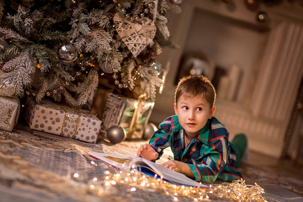 Chlapec leží na podlaze u zdobeného vánočního stromku. Čtu si knihu s vánočními příběhy. Útulné zimní večery doma. Koncept vánočních a silvestrovských prázdnin doma. selektivní zaměření; obrysové světlo; mělká hloubka pole - Fotografie, Obrázek