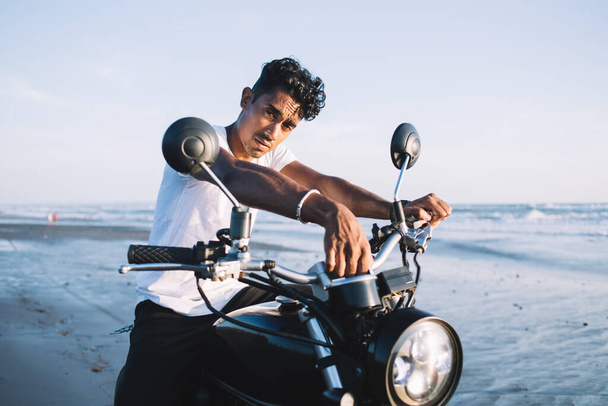 Handsome ethnic guy in white t shirt sitting on black motorcycle alone on empty beach at sunset and looking at camera - Foto, Imagen