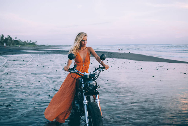 Привлекательная уверенная молодая женщина с длинными волосами в оранжевом платье, сидящая на мотоцикле и задумчиво глядящая на берег моря в сумерках - Фото, изображение