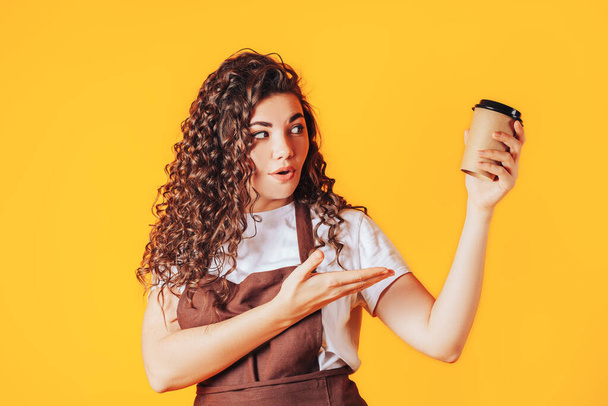 Γυναίκα που κρατάει ένα φλιτζάνι ζεστό καφέ στα χέρια της. Έκπληκτη γυναίκα δείχνει με το χέρι στο κύπελλο μιας χρήσης. Διαφήμιση σε καφετέρια. Θέση για το κείμενο σας - Φωτογραφία, εικόνα