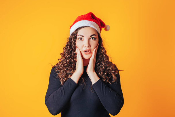 Beautiful woman with curly hair and santa hat on her head. The woman shows emotion of surprise and touched her face with her hands. Christmas mood. Space for text on yellow background. - Foto, Bild