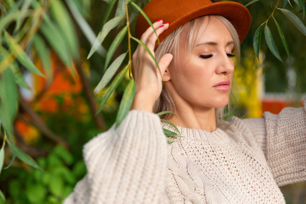 close up, young beautiful blonde woman in sweater, stands under branches and green leaves, holds felt hat fedora with her hand and closed her eyes. - Foto, immagini