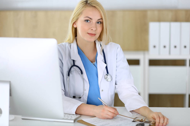 Doctor woman at work while sitting at the desk in hospital or clinic. Blonde cheerful physician filling up medical records form. Data and best service in medicine and healthcare - Фото, изображение