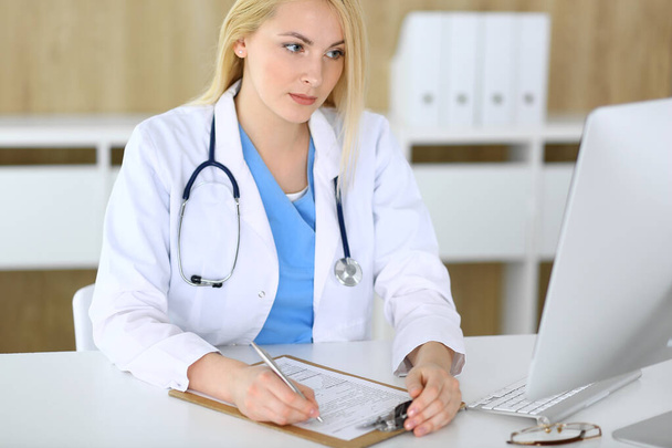 Doctor woman at work while sitting at the desk in hospital or clinic. Blonde cheerful physician filling up medical records form. Data and best service in medicine and healthcare - Foto, afbeelding