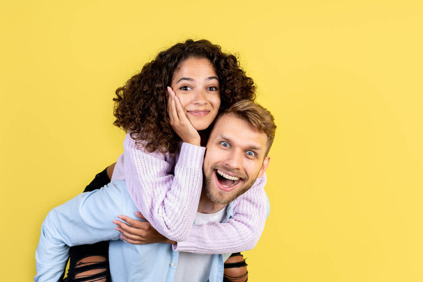 Portrait of excited man carry on the back happy african woman. Two mixed race people having fun together, standing on yellow background with copy space - Photo, Image