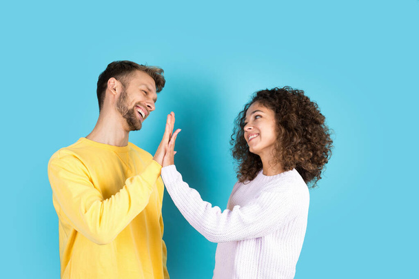Profile side view of happy man making greeting gesture give high five with smiling and young african woman. Two mixed race people clapping hands, standing on blue copy space background - Photo, Image