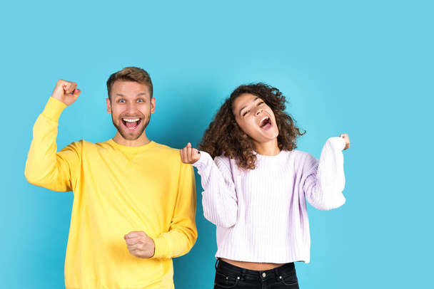 Celebration concept. Mixed race friends making rejoiced faces, smiling wide, screaming loud and raising fists up, standing together on blue background with copy space - Photo, Image