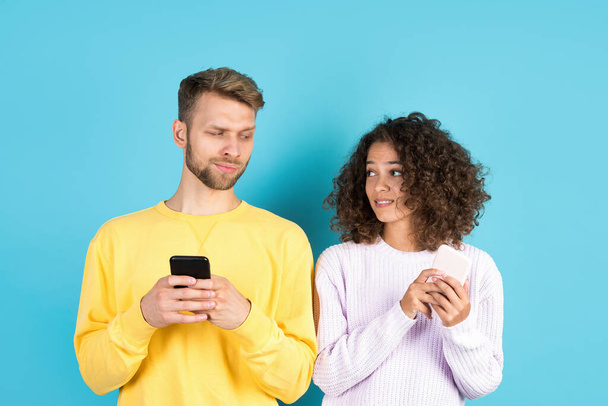 Afro american woman hiding secret at modern smartphone from man while they standing together on blue background with copy space. Interracial couple using online app on phone with wireless internet - Photo, Image