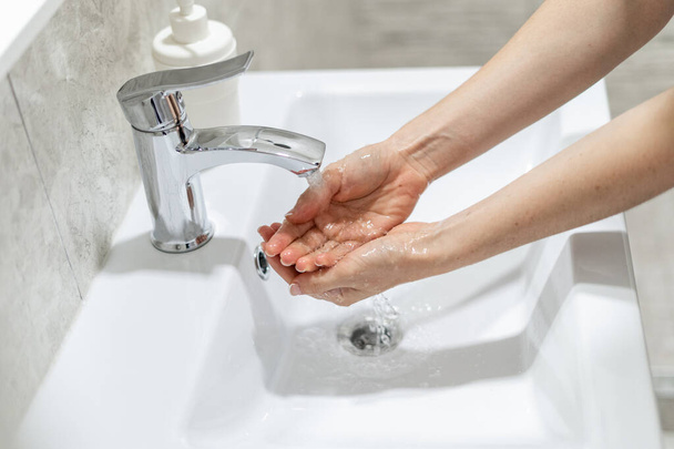 Concepto de prevención pandémica de la higiene y el coronavirus. Vista recortada de la mujer lavándose las manos bajo el agua. Muñeca de retención femenina sobre fregadero de cerámica en el baño - Foto, imagen