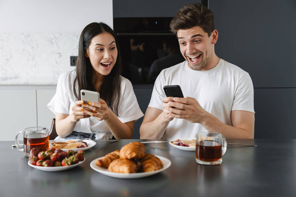Πορτρέτο ενός ευτυχισμένου νεαρού ζευγαριού που χρησιμοποιεί κινητά τηλέφωνα ενώ παίρνει νόστιμο πρωινό στο τραπέζι σε μια κουζίνα - Φωτογραφία, εικόνα
