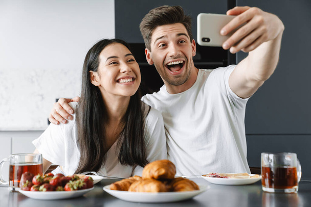 Young couple eating breakfast in the kitchen and taking selfie - 写真・画像