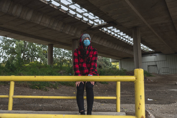 Young depressed homeless girl or woman with hood and protective medicine mask sitting outdoor under the bridge scared from coronavirus virus or COVID-19 feeling abandoned and anxious selective focus - Foto, imagen