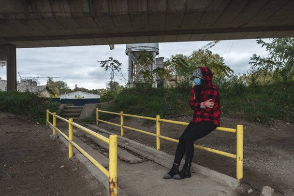 Young depressed homeless girl or woman with hood and protective medicine mask sitting outdoor under the bridge scared from coronavirus virus or COVID-19 feeling abandoned and anxious selective focus - Foto, imagen