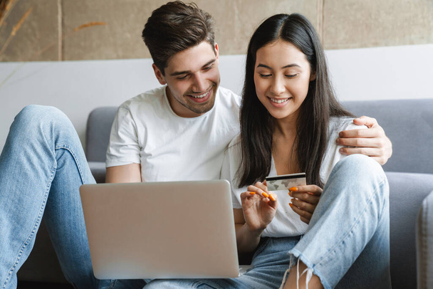 Photo of a cheerful young loving couple at home on floor near sofa using laptop computer and holding credit card - Φωτογραφία, εικόνα